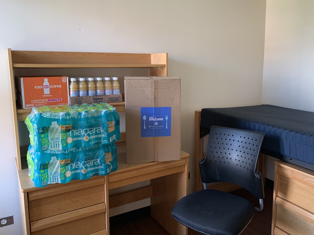 A dorm room that has a Welcome Box along with a supply of water, juice, and coffee sitting on a desk.