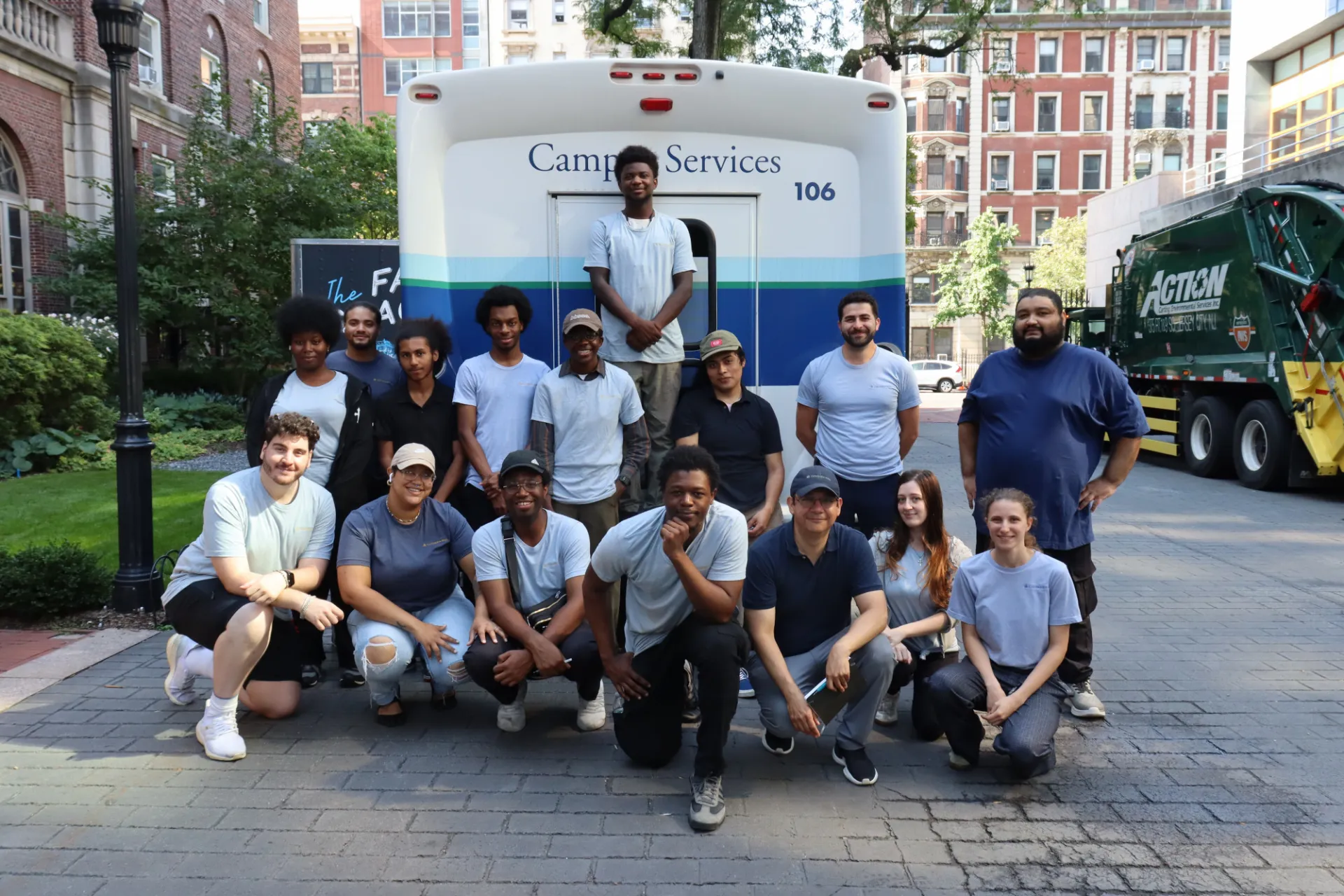 A group of Campus Services employees pose together in front of a Campus Services truck on a campus walkway. The team is arranged in several rows, with campus buildings, trees, and a service vehicle visible in the background.