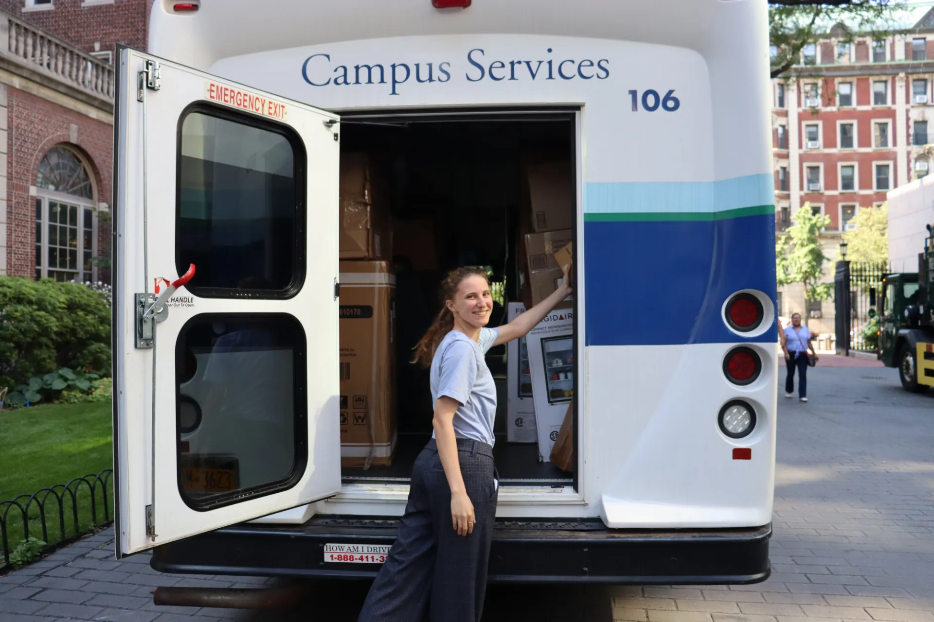 Campus Services truck parked on a campus walkway with the rear door open, revealing boxes and equipment inside. A staff member stands at the back of the vehicle while unloading supplies.