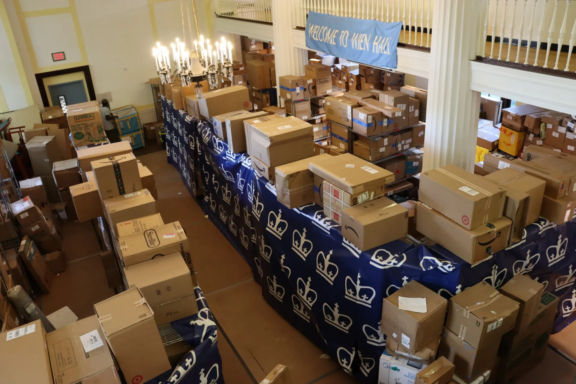 Large residence hall package distribution area filled with labeled boxes and parcels organized on tables beneath a “Welcome to Wien Hall” banner during move-in season.